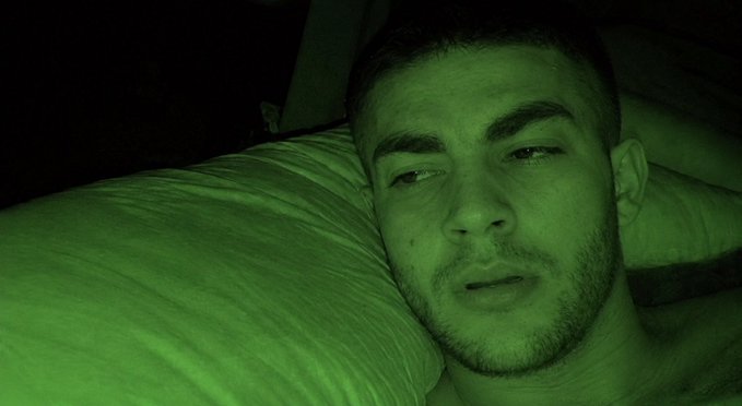A man lies in bed in a dimly lit room with a greenish tint, possibly from night vision or low lighting.