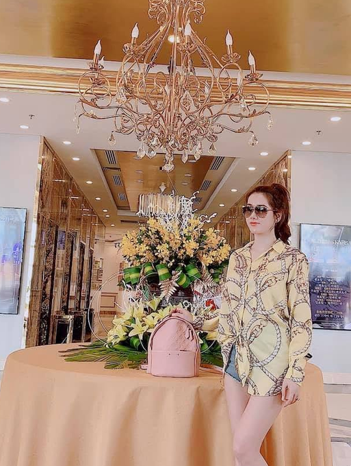 A woman wearing sunglasses and a yellow patterned shirt poses indoors next to a table featuring a pink backpack and floral arrangements under an ornate chandelier.