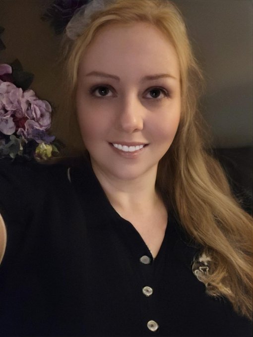 A selfie of a smiling young woman with long blonde hair, wearing a black button-up shirt, with purple flowers visible in the background.