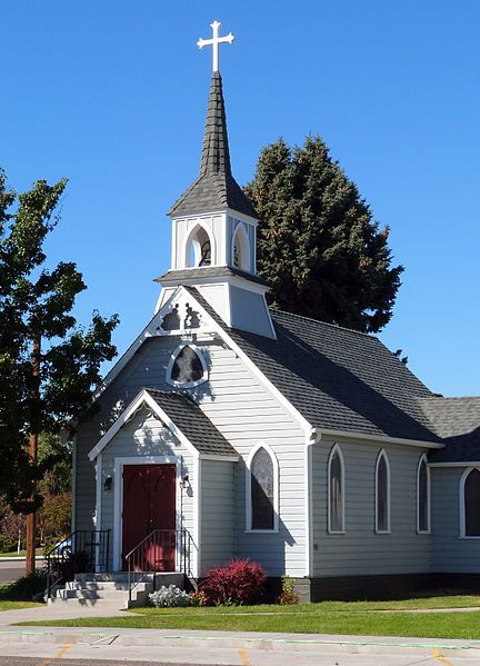 The image features a small white church with a tall steeple and cross at its peak. The church has a red door, which stands out against the white exterior of the building. In front of the church, there are some bushes that add greenery to the scene. A car is parked in front of the church, indicating that it might be a popular spot for visitors or locals alike.