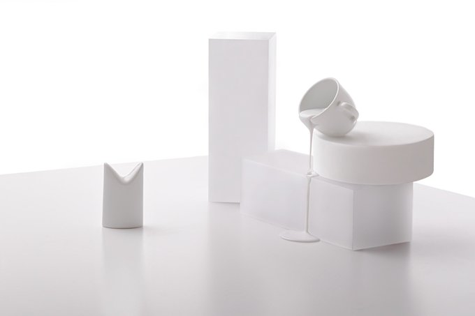 A minimalist arrangement of white geometric objects, including a tipped cylinder spilling liquid onto geometric blocks of various shapes, against a white background.