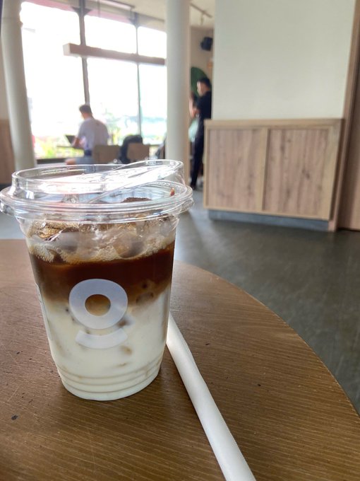 An iced coffee drink in a clear plastic cup with a domed lid and a white paper straw, sitting on a wooden table inside a bright cafe with people visible in the background.