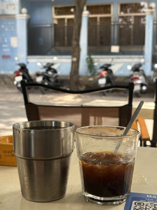 A metal tumbler and a glass of iced coffee with a spoon sit on a table at a street-side café, with motorbikes parked across the way in front of a blue building.