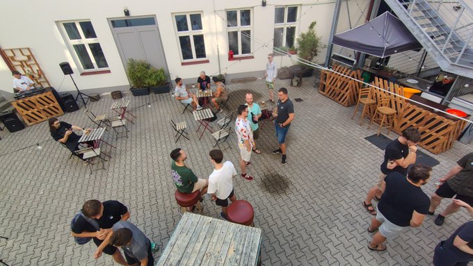 An aerial view of a courtyard gathering where people are mingling around tables, chairs, and a large umbrella, with a building featuring tall windows in the background.