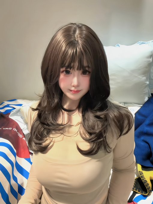 A woman with long, wavy dark hair and blunt bangs poses indoors wearing a fitted beige long-sleeve top.