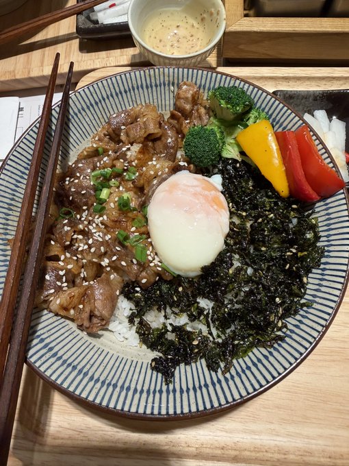 A Japanese rice bowl dish topped with teriyaki beef, a soft-boiled egg, sesame seeds, broccoli, and bell peppers, served with seaweed strips and a side of creamy sauce.