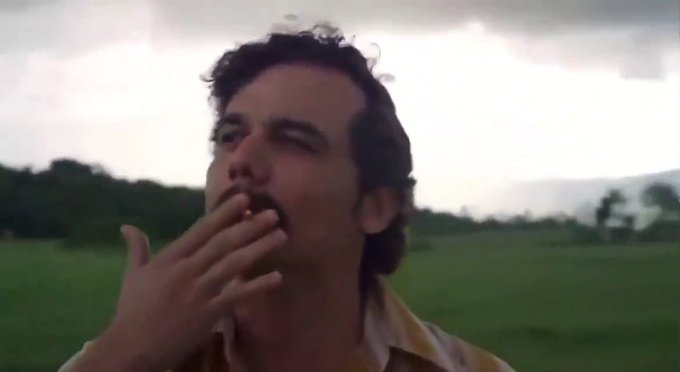 A close-up of a man with dark hair and a mustache smoking a cigarette in a green field, with a cloudy sky in the background.