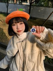 Portrait-style photo of a young woman wearing an orange beret and a cream cable-knit sweater, holding up a small blue gear-shaped object near her face in an outdoor setting.