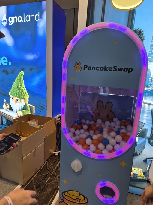 A blue and pink neon-lit arcade claw machine branded with 'PancakeSwap' filled with orange, blue, and white balls, displayed next to a Lego gnome figure.
