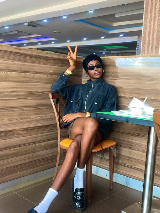 A woman in a dark denim outfit and sunglasses sitting at a restaurant booth, throwing up a peace sign.