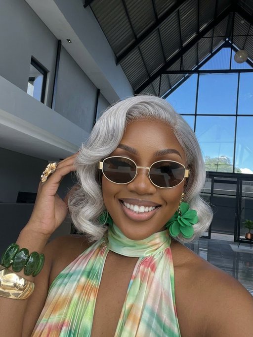 A smiling woman poses outdoors wearing silver curled hair, narrow oval sunglasses, green flower earrings, a pastel scarf, green bracelets, and gold rings.