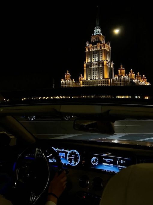 A nighttime view from inside a car showing the dashboard and steering wheel, looking out at an illuminated, ornate high-rise building with the moon visible in the sky.