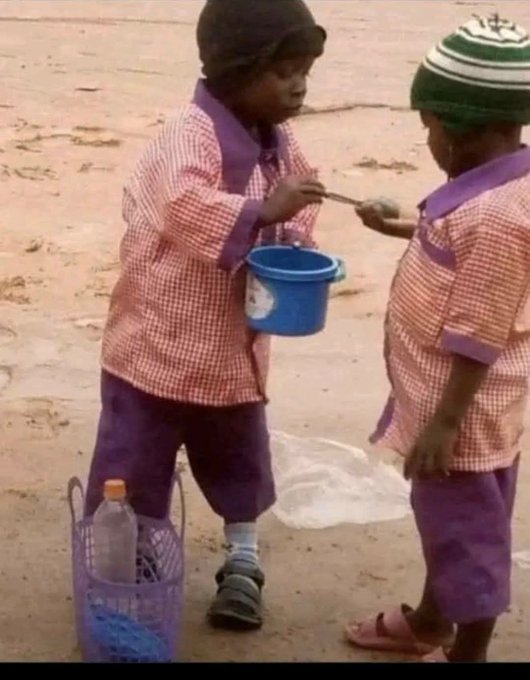 A low-resolution photo of two young children in matching pink and purple school uniforms sharing a blue bucket, with a wire basket and plastic bottle nearby on a sandy ground.