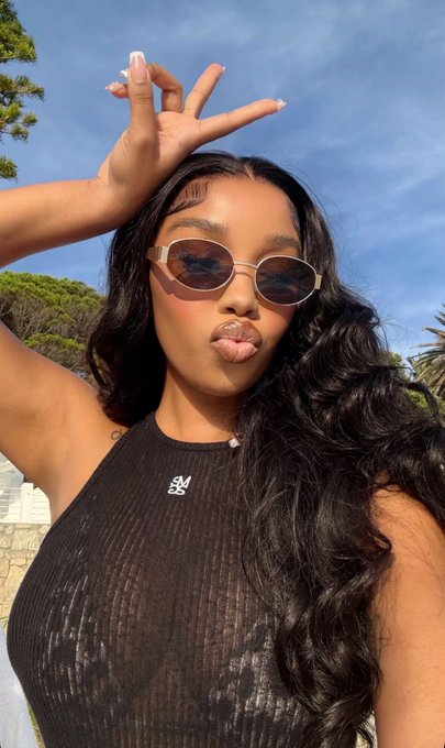 A young woman with long, wavy black hair poses playfully outdoors against a clear blue sky, making a peace sign above her head and puckering her lips. She wears brown-tinted oval sunglasses and a black mesh halter top with a small white logo resembling "SM," perfectly matching her relaxed, fun "happy sunday crypto friends" vibe.
