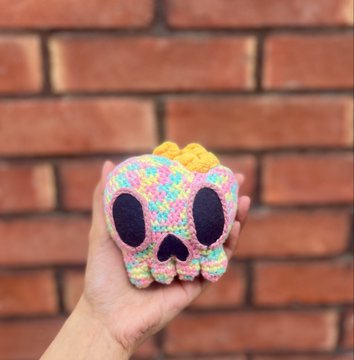 A hand holding a small, colorful crocheted skull with yellow accents on top, displayed against a red brick wall background.