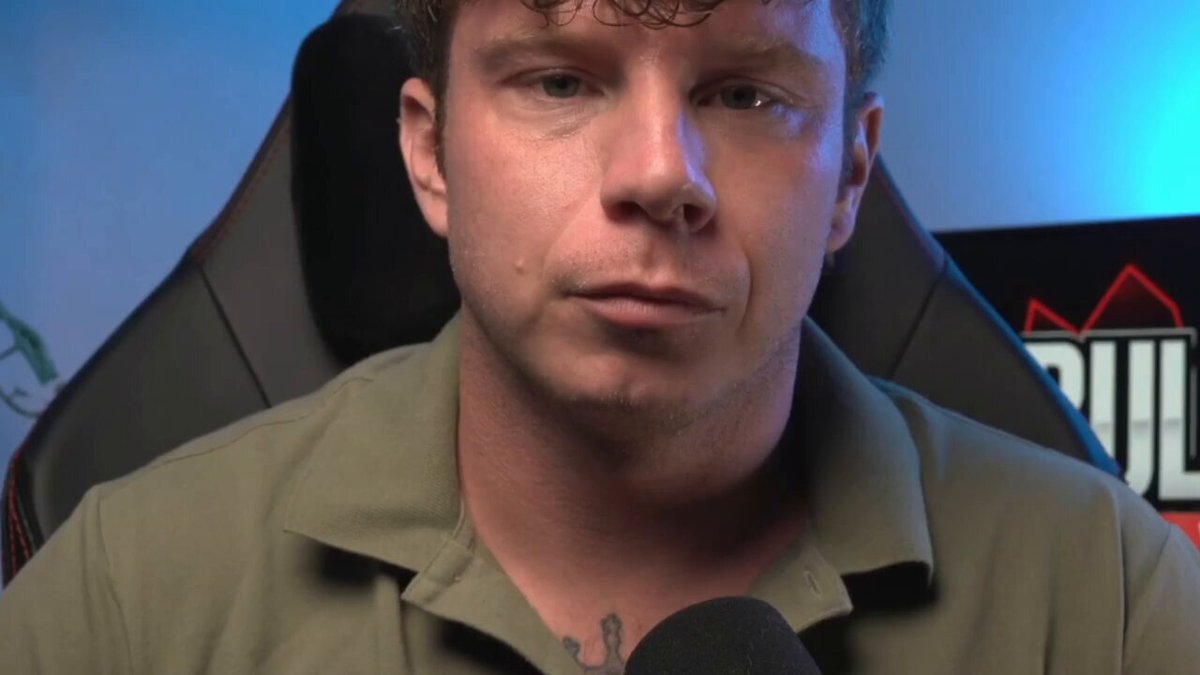 The image shows a man wearing a green shirt, sitting in a chair with his face turned towards the camera. He has tattoos visible on his neck and chest area. The background of the image features a blue wall with a poster hanging on it that says "Ripple XRP THEY HID IT! Lloyd Blankfein CONFIRMED What'S About To Hit The Banks!". This suggests that the man might be participating in an online gaming event or discussion related to Ripple XRP, as indicated by the text on the poster.