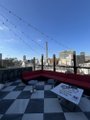 A small rooftop terrace with checkered flooring overlooks a city skyline under a clear blue sky, with a red and white umbrella and string lights adding a casual ambiance. The image complements the post about settling drinks with friends after a study session using Lightning payment.