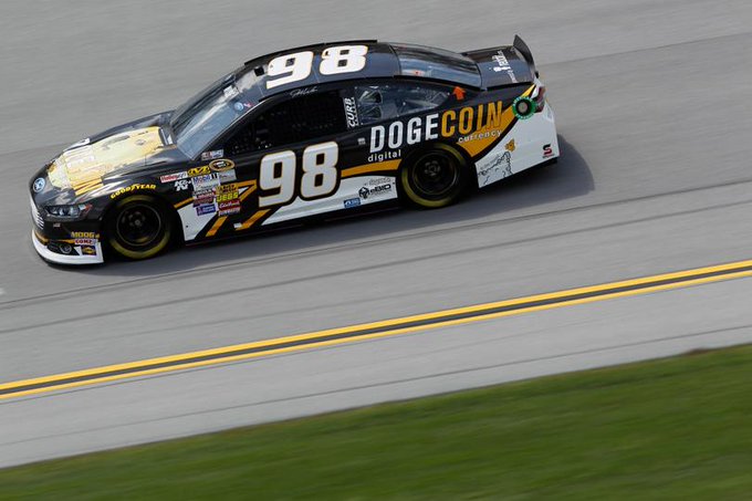 A NASCAR race car #98 featuring Dogecoin/DogeCoin Digital branding in black, white, and yellow color scheme racing on a track.