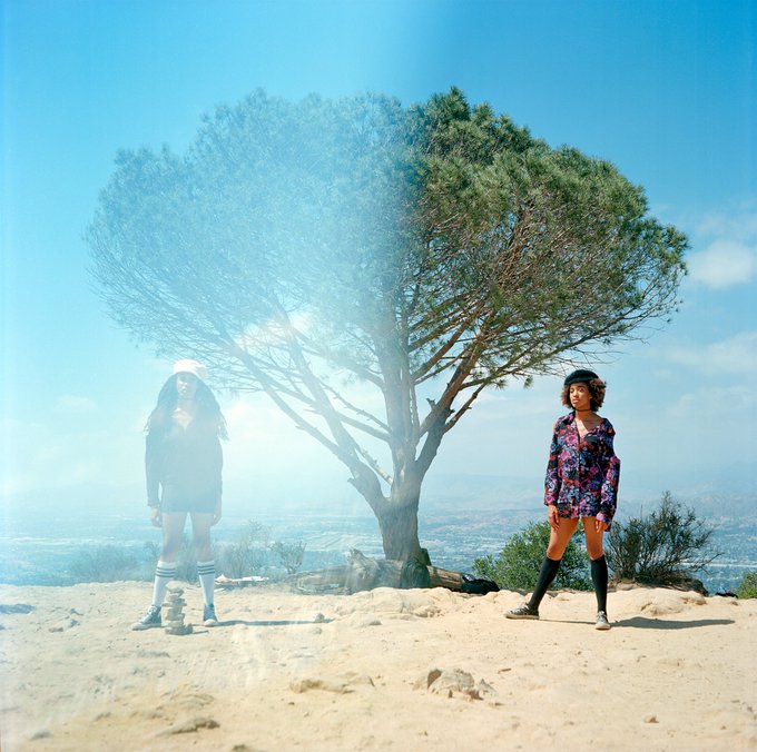 Two young women standing on a sandy overlook beneath a large tree, with one figure appearing as a translucent, ghostly overlay on the left side of the image.
