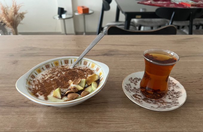 A bowl of creamy dessert topped with cinnamon, almonds, and fruit, served next to a traditional tulip-shaped glass of tea on a wooden table.