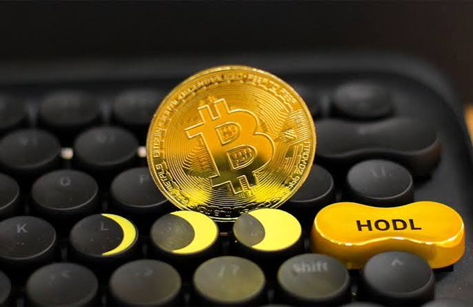 A golden Bitcoin coin rests on a black keyboard next to a yellow key cap labeled "HODL", symbolizing cryptocurrency investment.