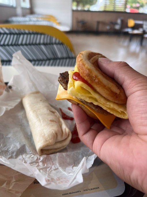 A hand holds a partially eaten McDonald's McMuffin-style breakfast sandwich with visible cheese and ketchup, alongside a breakfast burrito on wrapper paper showing "©2025 McDonald's VOX1BURGER" text, inside what appears to be a McDonald's restaurant.