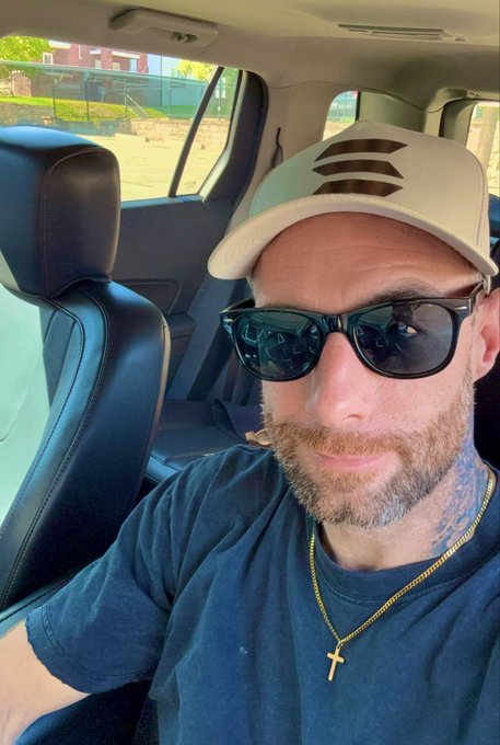 A close-up selfie of a man in a car wearing sunglasses, a white cap, a black shirt, and a gold cross necklace.