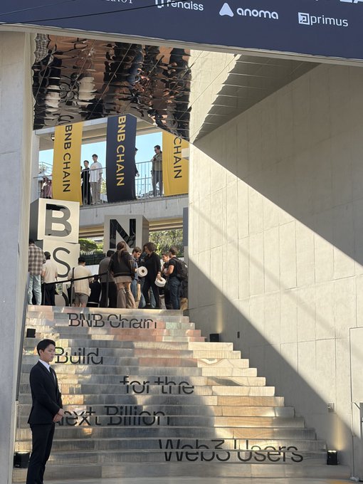 An outdoor venue features stairs with projected text reading 'BNB Chain: Build for the Next Billion Web3 Users', with yellow 'BNB CHAIN' banners hanging above and sponsor names like Anoma, Primus, and others on a top banner.