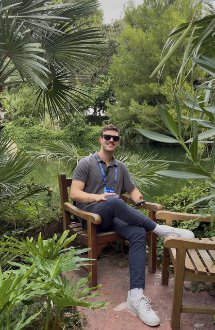 A man wearing sunglasses, a gray polo shirt, and a lanyard sits on a wooden bench in a lush garden setting surrounded by palm trees.