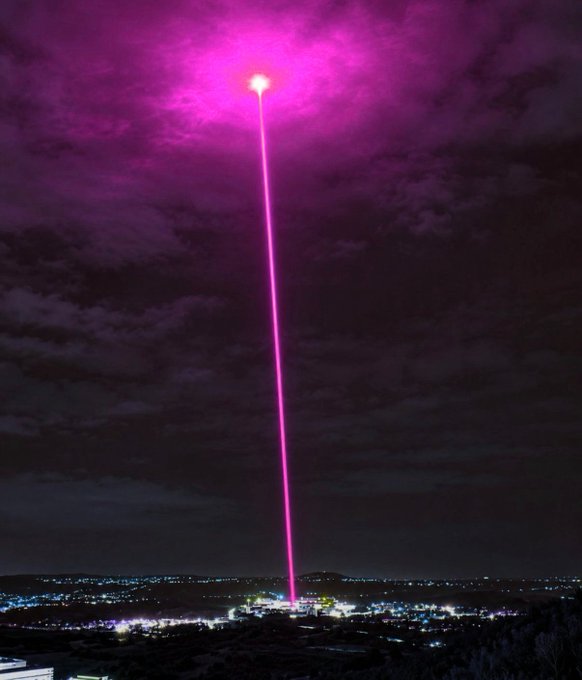 A nighttime cityscape featuring a dramatic bright pink/magenta laser beam shooting straight up into the cloudy night sky from a city below.