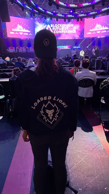 A person wearing a 'Loaded Lions' hoodie and beanie standing at a blockchain futurist conference, viewed from behind looking toward the stage.