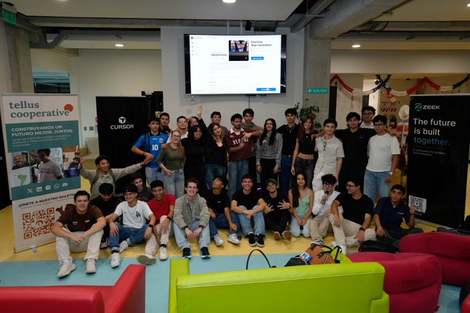 A large group of students or participants posing for a group photo at what appears to be a tech or cooperative event, with sponsor banners including 'Tellus cooperative,' 'Cursor,' and 'Zeek' visible, in a colorful lounge space.