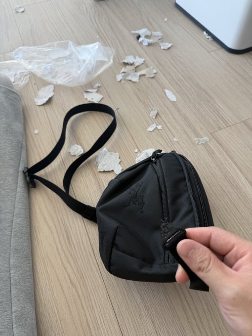 A hand holding a black sling bag on a wooden floor scattered with torn paper and a plastic bag.