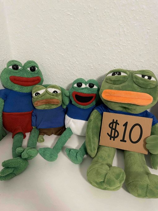 A group of four Pepe the Frog plush toys, with the largest one on the right holding a cardboard sign that says '$10'.