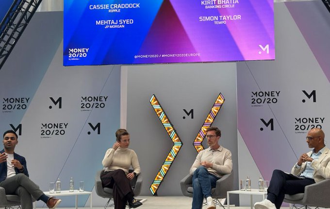 The image shows four people sitting around a table at an event, likely discussing business or finance topics. The panel consists of three men and one woman who are seated behind a large screen displaying information related to money and banking. One man appears to be speaking while the other two sit attentively in front of him. There is also a banner hanging above them that reads "MONEY 20:20".