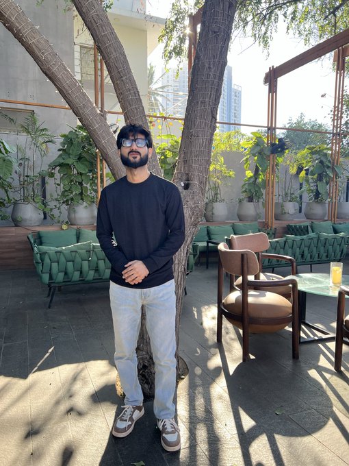 A young man with a beard, wearing sunglasses, a black long-sleeve shirt, and light jeans, stands leaning against a tree in an outdoor patio area with green sofas and lounge chairs.