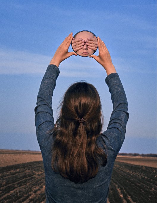 An artistic photograph of a woman seen from behind holding a round mirror up to the sky, reflecting a face with hands covering its eyes.