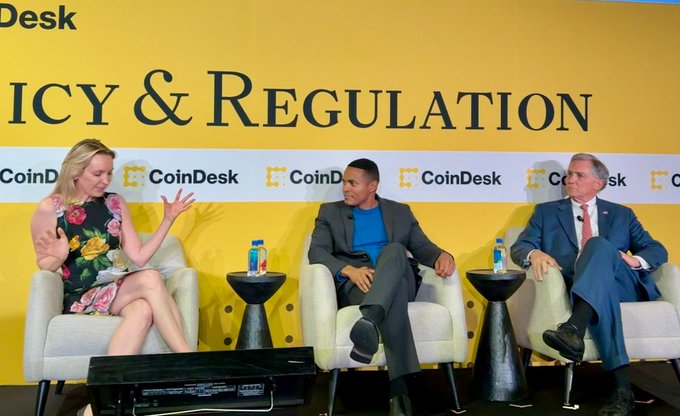 Three people participate in a panel discussion at a CoinDesk event titled "Policy & Regulation," seated on a stage with a bright yellow backdrop.