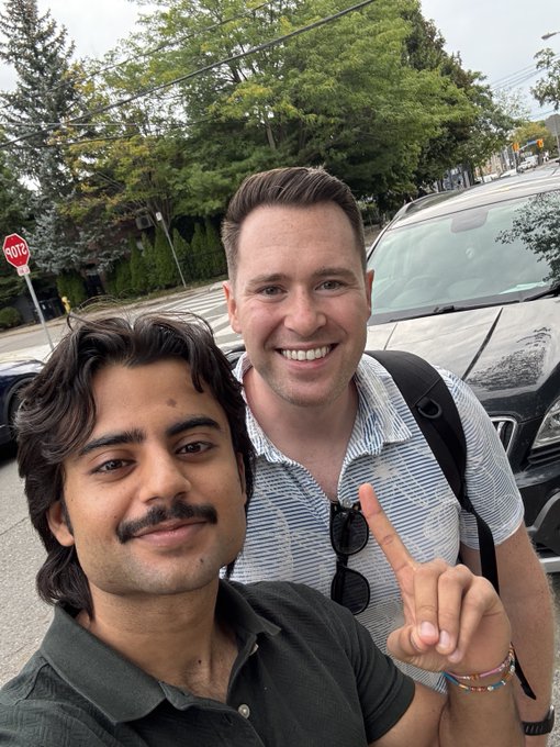 A selfie of two smiling men on a city street; the man on the right wears a striped shirt and makes a peace sign, while the other wears a dark polo.
