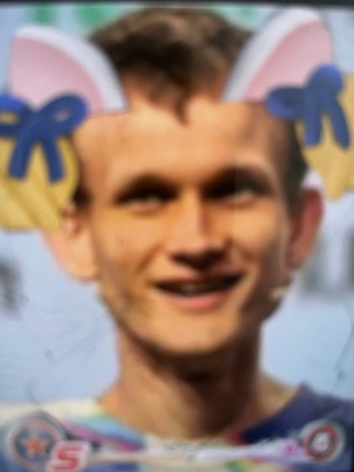 A young man with short brown hair smiles while wearing a digital filter that adds pink bunny ears with blue bows and a colorful patterned shirt overlay.
