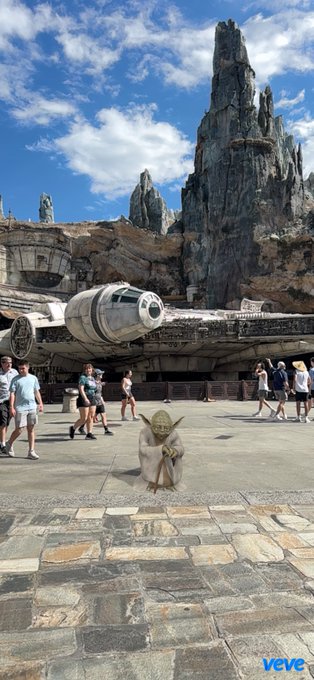 A Yoda character is edited into a photo at what appears to be Star Wars: Galaxy's Edge at Disneyland, sitting on the stone ground near the Millennium Falcon with rocky spires in the background.