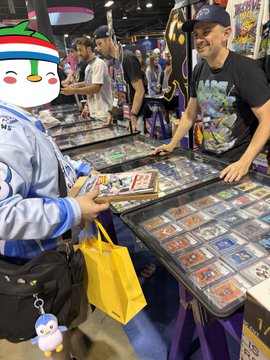 A photograph taken at what appears to be a trading card or collectibles convention, showing a person browsing cards at a booth while another attendee in a snowman hoodie looks on.