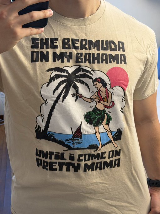 A mirror selfie of a person wearing a beige t-shirt featuring a hula dancer graphic and the text 'SHE BERMUDA ON MY BAHAMA UNTIL I COME ON PRETTY MAMA'.