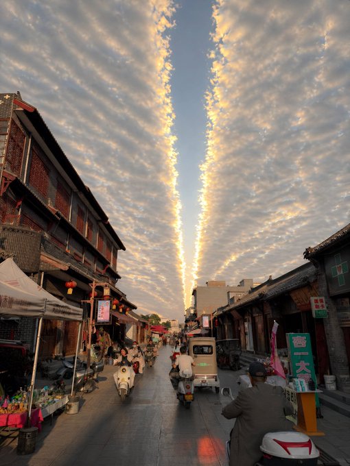 A scenic view of a traditional Chinese street at sunset, featuring distinctive cloud formations, historic buildings with red lanterns, and people on scooters.