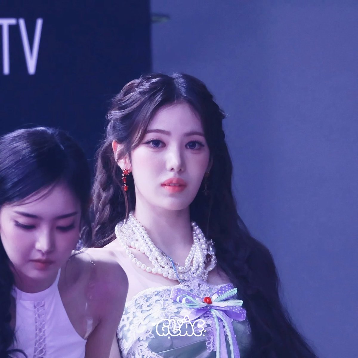 A K-pop idol performing on stage wearing an elegant white and pastel dress with elaborate pearl necklaces, with another performer visible in the background.