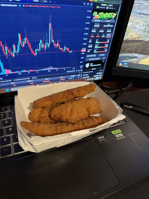 Fried chicken tenders sit in a takeout box on a laptop keyboard, with a cryptocurrency price chart displayed on the monitor in the background showing a $1,439.32 ticker.