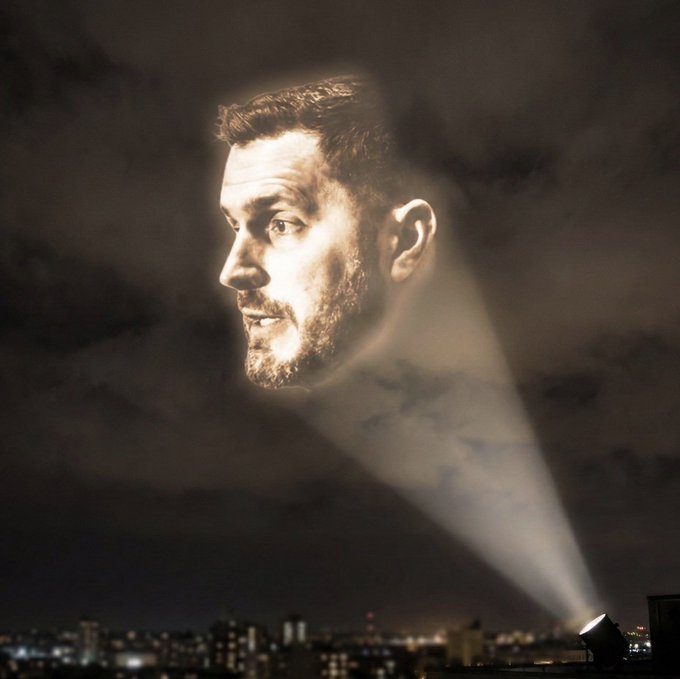A surreal composite image showing a bearded man's profile illuminated against a dark night sky, with a spotlight from the ground projecting his image up into the clouds above a city skyline.