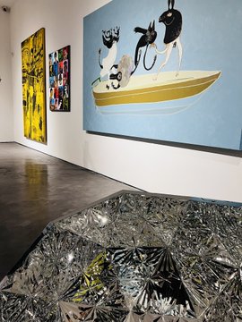 A person stands in an art gallery admiring contemporary works, including a whimsical painting of stylized figures on a yellow boat against a blue background, alongside vibrant abstract canvases and a textured metallic floor installation—perfectly capturing the peaceful, reflective atmosphere of being immersed in art.