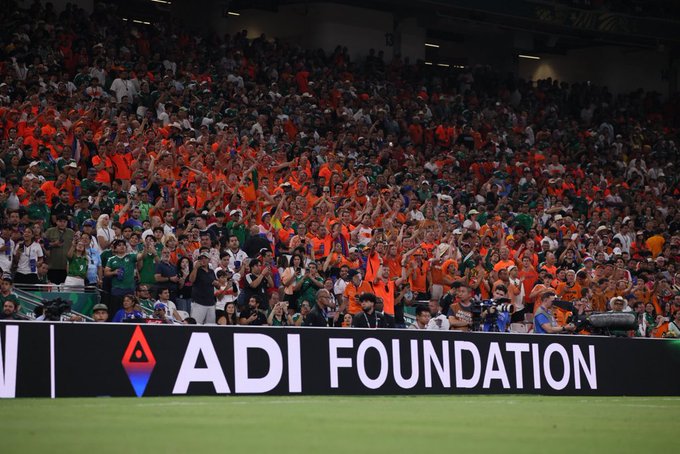 A lively stadium crowd, many wearing orange, cheers enthusiastically from the stands behind a prominent black barrier displaying the white "ADI FOUNDATION" logo with its distinctive red and blue triangular emblem. The image visually connects to the post by highlighting the ADI Foundation's prominent presence at the World Cup, reinforcing its status as a top global partner alongside major brands.