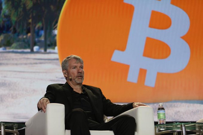 A gray-haired, bearded man in a black suit sits on a white armchair on stage, with a large Bitcoin logo displayed on a screen behind him, relating to the post about Saylor's Strategy selling $100 million in Bitcoin amid ongoing crypto struggles.